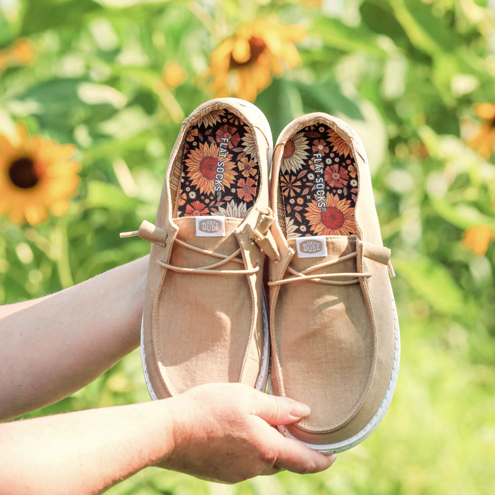 Pair of tan shoes with floral designs on Harvest Hue FLAT SOCKS, held against a sunflower field #size_small-up-to-women-s-11-men-s-10