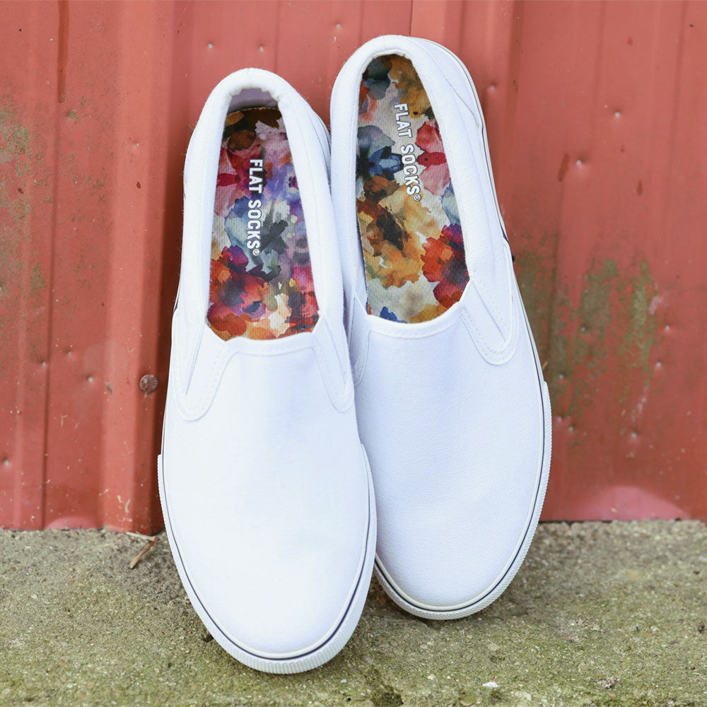 White slip-on shoes with colorful Watercolor Wildflower FLAT SOCKS inside, sitting against a red metal wall. #size_small-up-to-women-s-11-men-s-10