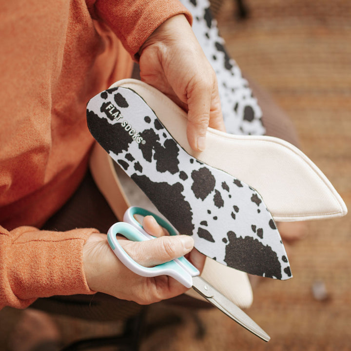 Woman in orange long-sleeved shirt cutting Black Cow Print FLAT SOCKS to size and shape of narrow-toed flat shoe #size_small-up-to-women-s-11-men-s-10