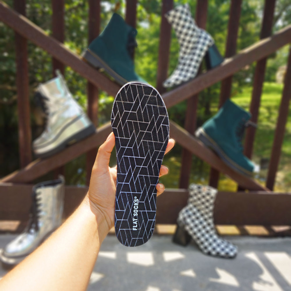 Person holding up Black FLAT SOCKS, different pairs of boots in the background #size_small-up-to-women-s-11-men-s-10