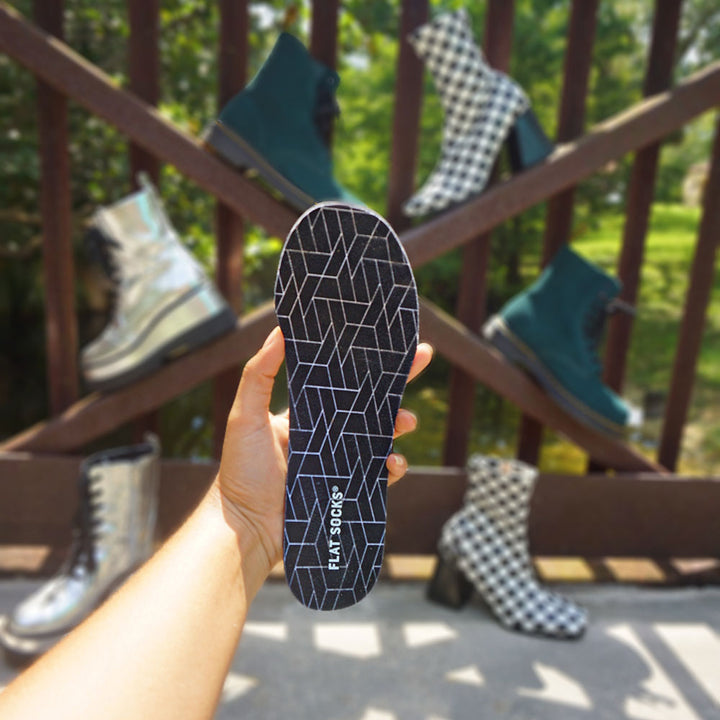 Person holding up Black FLAT SOCKS, different pairs of boots in the background #size_small-up-to-women-s-11-men-s-10