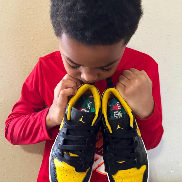 Little boy in red shirt holding up a pair of black and yellow sneakers with Dinosaur FLAT SOCKS Kids inside #size_8-toddler-to-4-kid-s