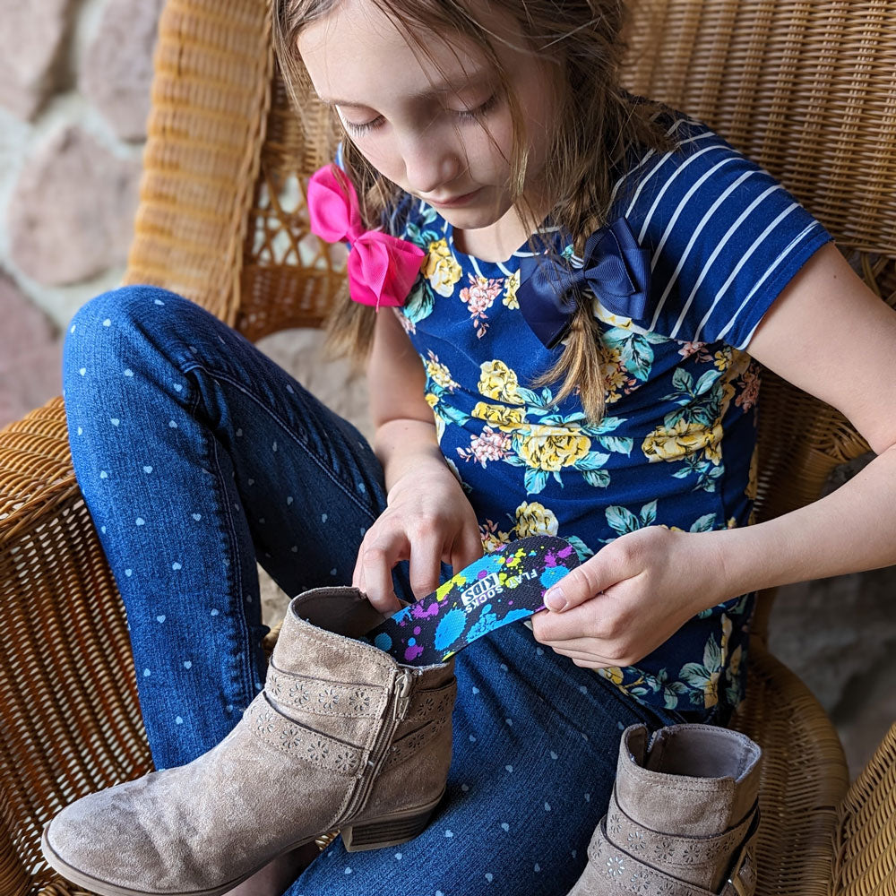 Little girl sitting on front porch in wicker chair placing Paint Splatter FLAT SOCKS Kids into beige suede booties #size_8-toddler-to-4-kid-s