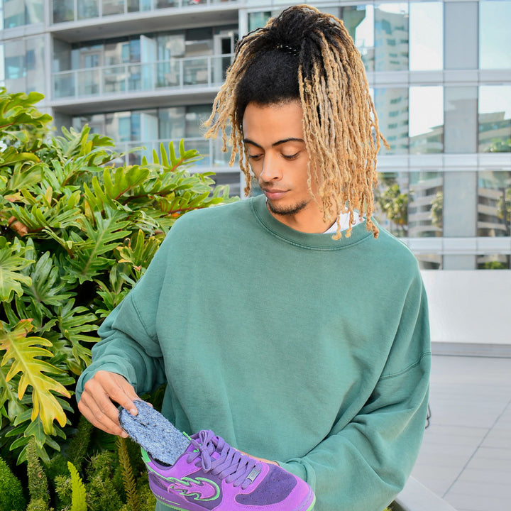 Man standing outside on balcony with green plants next to him while holding a pair of purple and green tennis shoes with Koala Faux Fur FLAT SOCKS inside #size_small-up-to-women-s-11-men-s-10