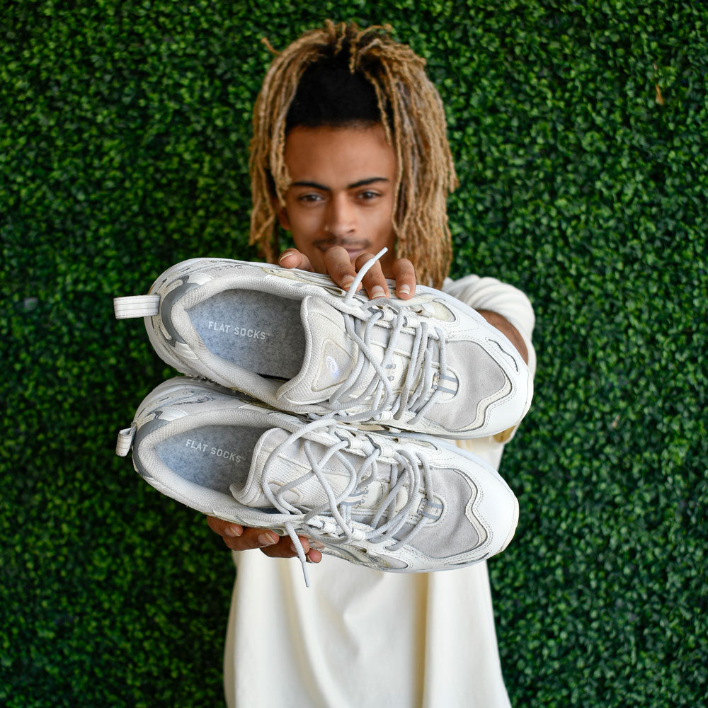 Man standing in front of green bush background while holding up a pair of white sneakers with Light Heather Gray FLAT SOCKS inside #size_small-up-to-women-s-11-men-s-10