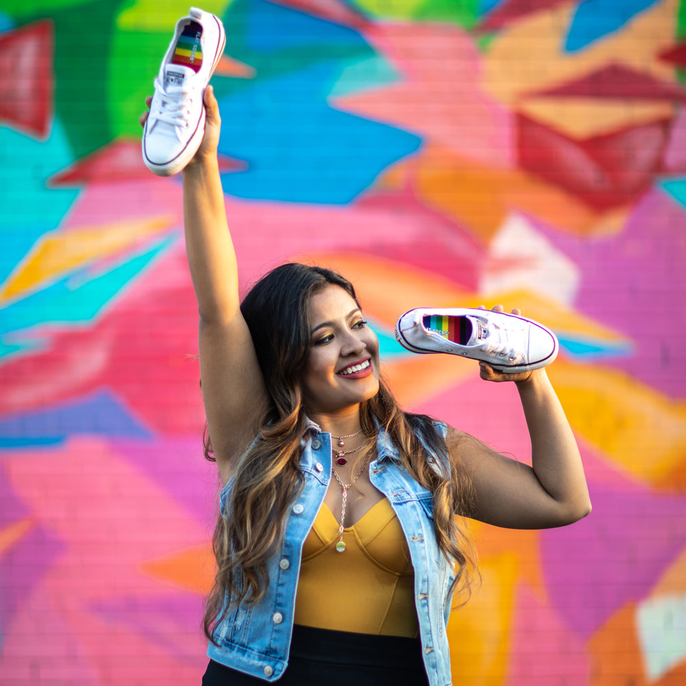 Woman standing in front of colorful mural while holding up white Converse shoes with Rainbow FLAT SOCKS inside #size_small-up-to-women-s-11-men-s-10