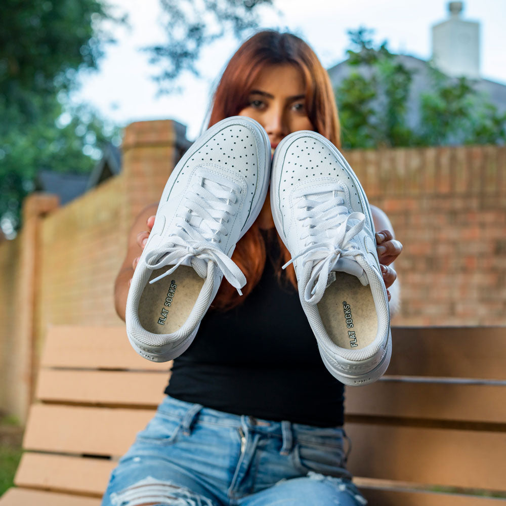 Woman sitting outside of a brick fence on a bench while holding up a pair of white tennis shoes with Sand FLAT SOCKS inside #size_small-up-to-women-s-11-men-s-10