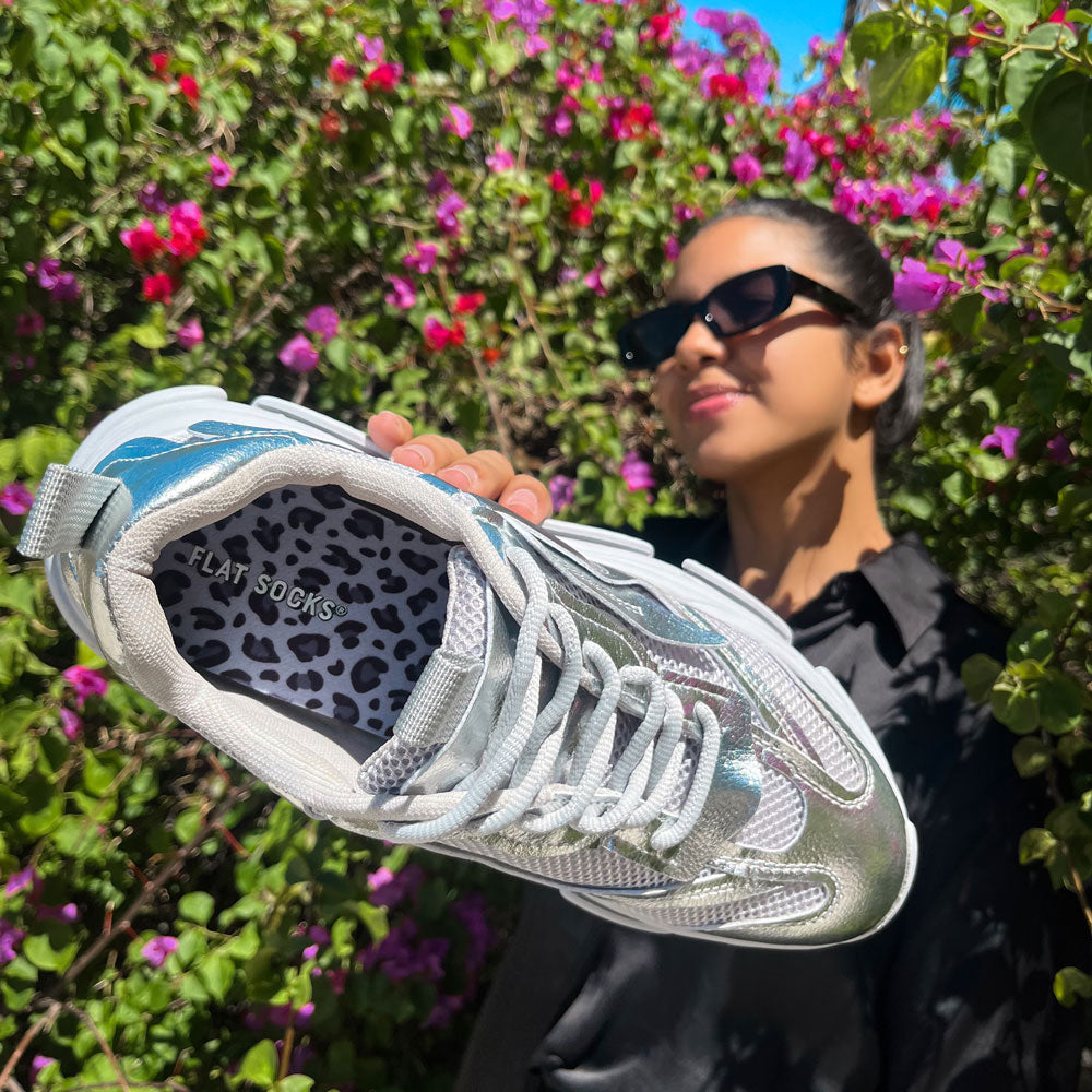 Woman wearing black sunglasses and standing in front of pink and red flowering bush while holding up a silver metallic colored sneaker with Snow Leopard FLAT SOCKS inside #size_small-up-to-women-s-11-men-s-10